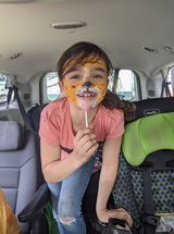 A little girl with her face painted like a tiger is sitting in the back seat of a car.