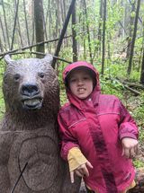 A little girl is standing next to a bear statue in the woods.