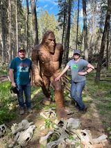 A man and a woman are standing next to a statue of a bigfoot in the woods.