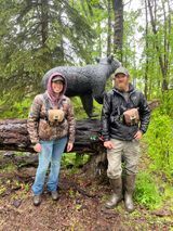 A man and a woman are standing next to a statue of a bear in the woods.
