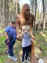 A woman and a child are standing next to a statue of a bigfoot in the woods.