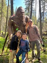 A man and two children are standing in front of a statue of a bigfoot in the woods.