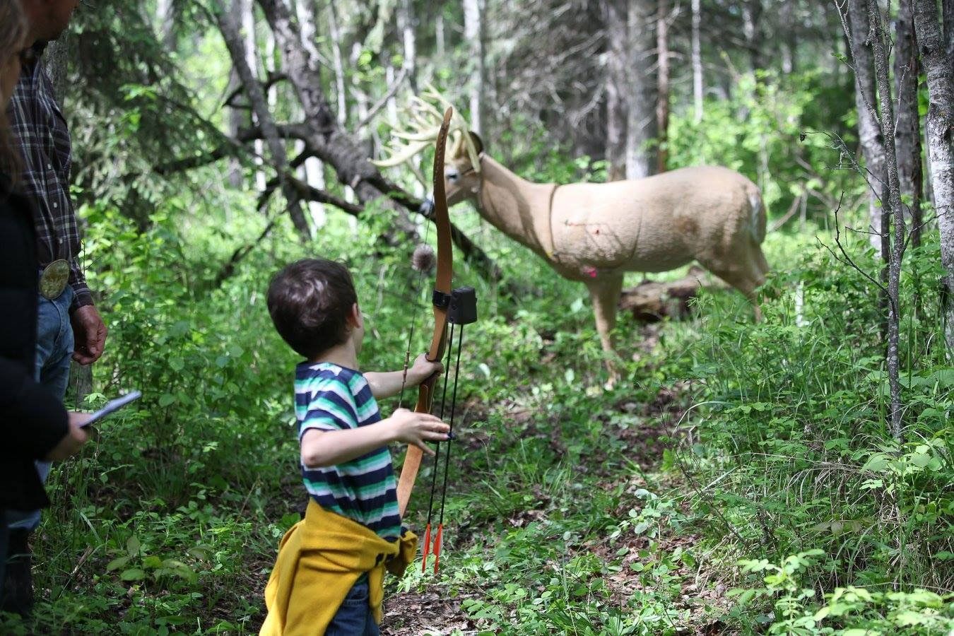 A young boy is aiming a bow at a deer in the woods.