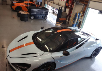 White McLaren sports car with orange stripe parked in a garage; another orange car in background.