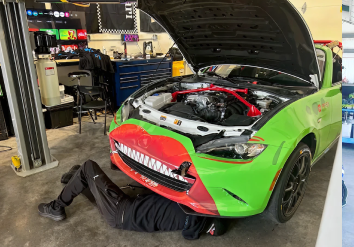 Mechanic under a green and red car with its hood open in a garage.