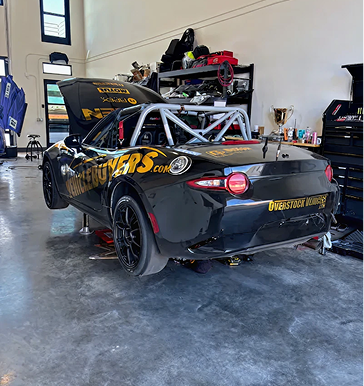 Black Mazda Miata race car, jacked up, in a garage. Roll cage visible, doors open. Gold lettering.
