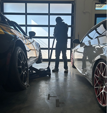 Person in garage with a floor jack between two cars. Sunlight, concrete floor, garage door visible.