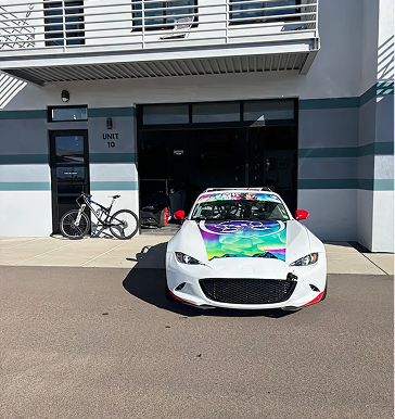 White sports car with colorful hood wrap parked in front of a building with a bike.