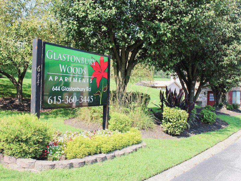 Sign for Glastonbury Woods Apartments, green with red leaf, shrubs, flowers, and trees.