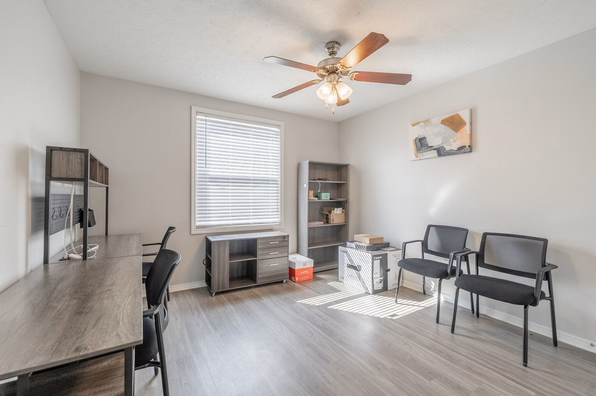Office room with desk, chairs, shelving, window, ceiling fan, and art. Gray walls, wood-look floor at  Glastonbury Woods Apartments.