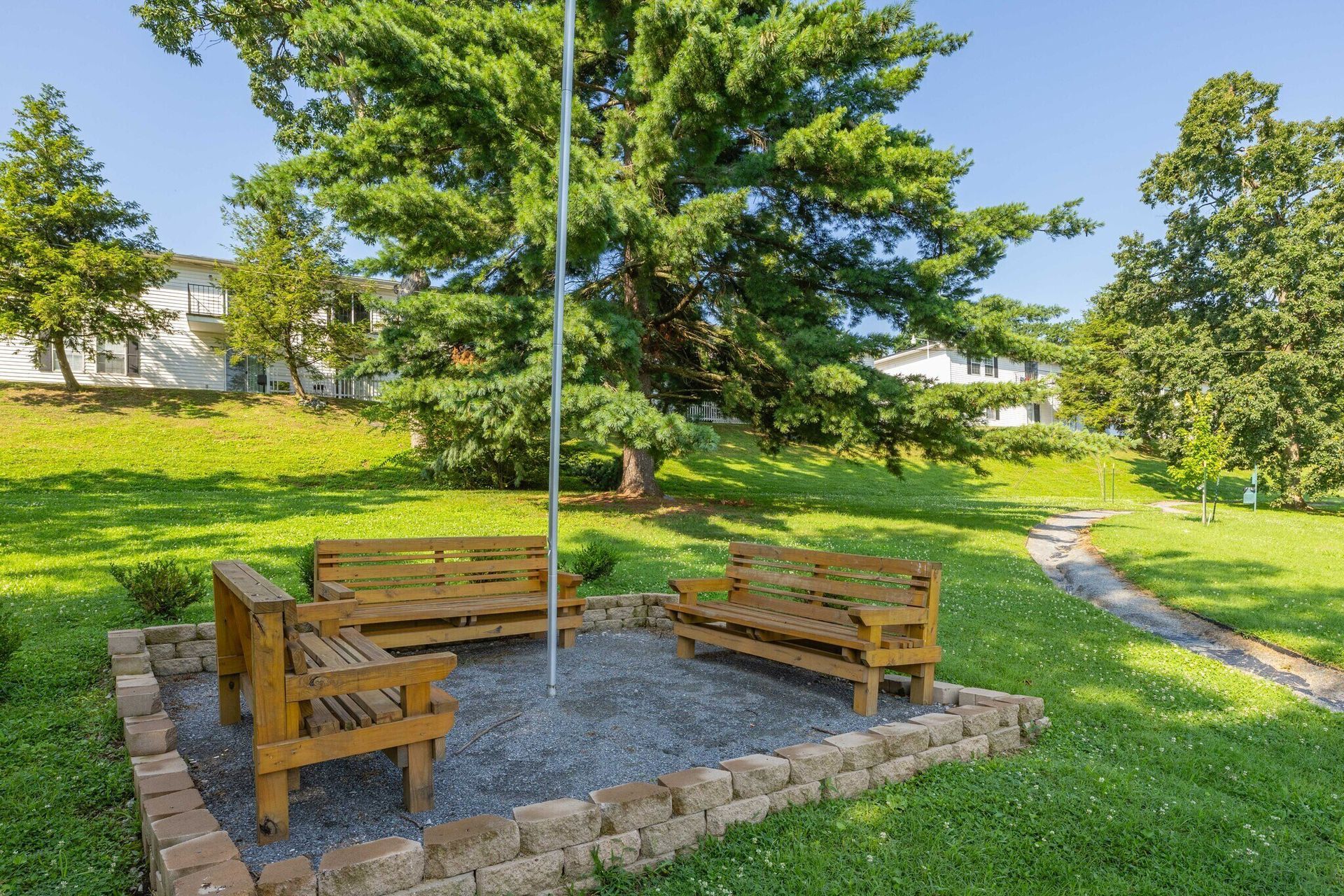 Three wooden benches around a flagpole on a gravel bed within a brick-lined area; grassy yard, large tree.