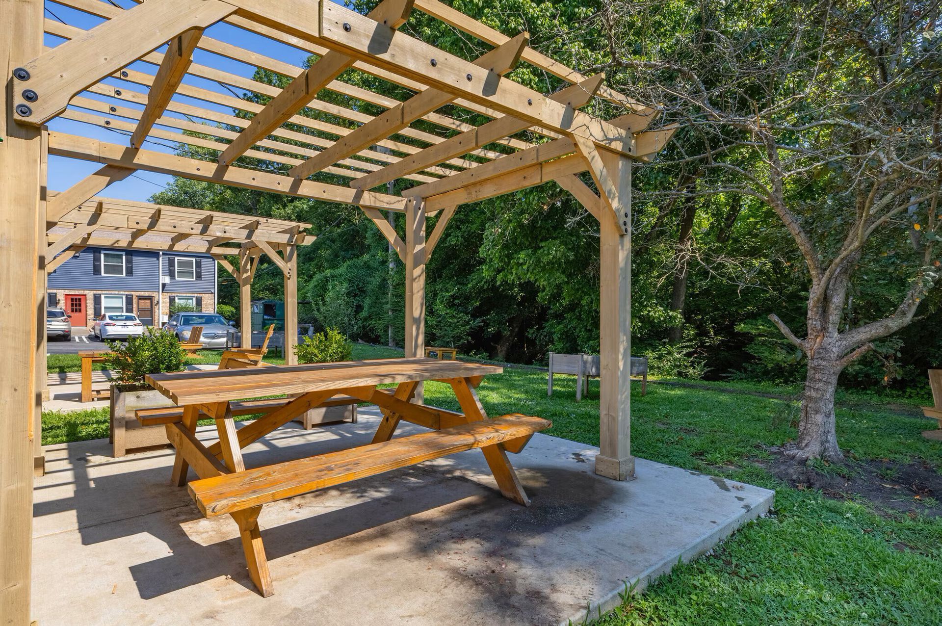 Wooden picnic table under a pergola on a concrete patio, with green trees and buildings in the background at  Glastonbury Woods Apartments.