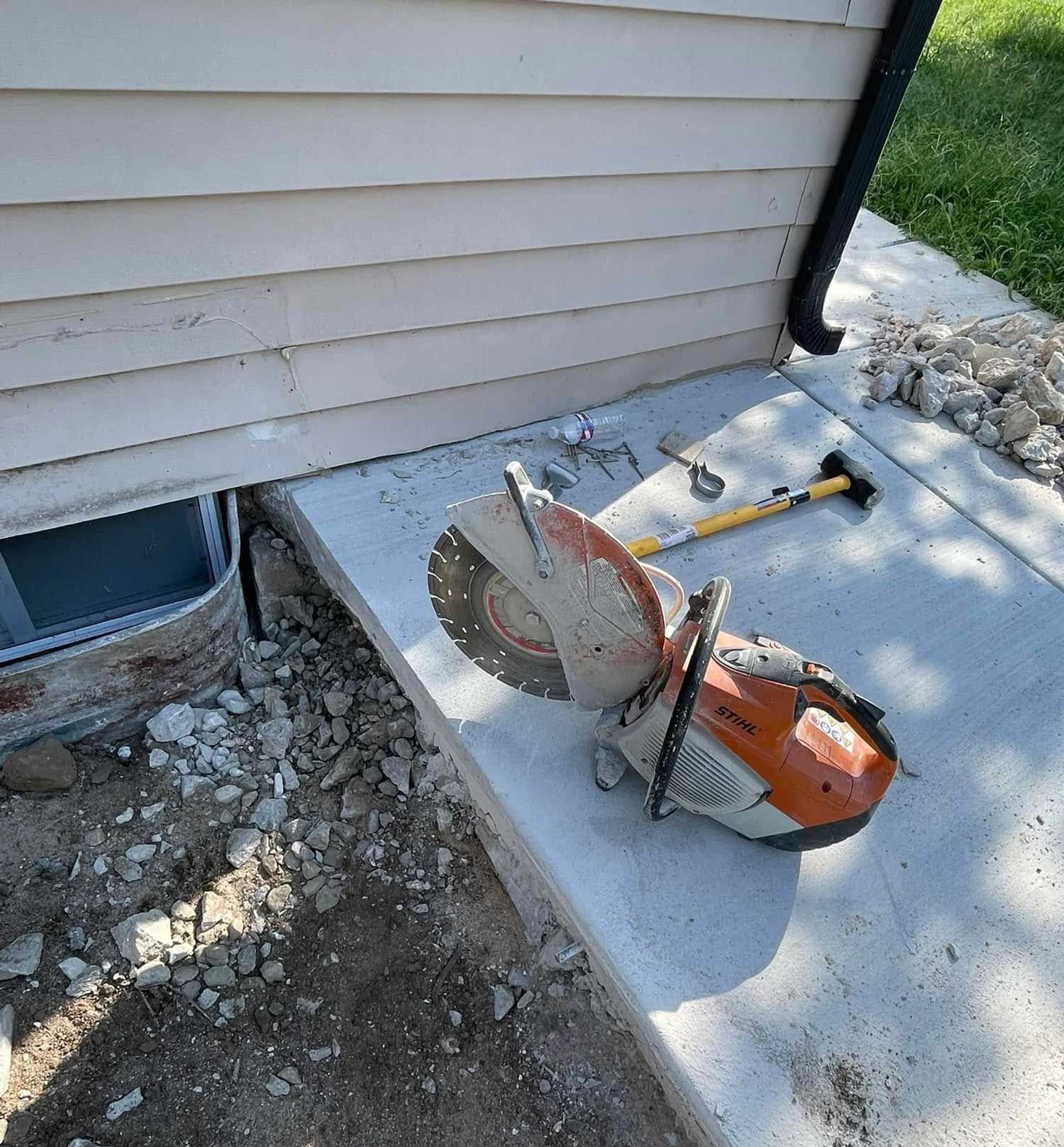 A power saw on a concrete surface next to a house; debris on the ground.