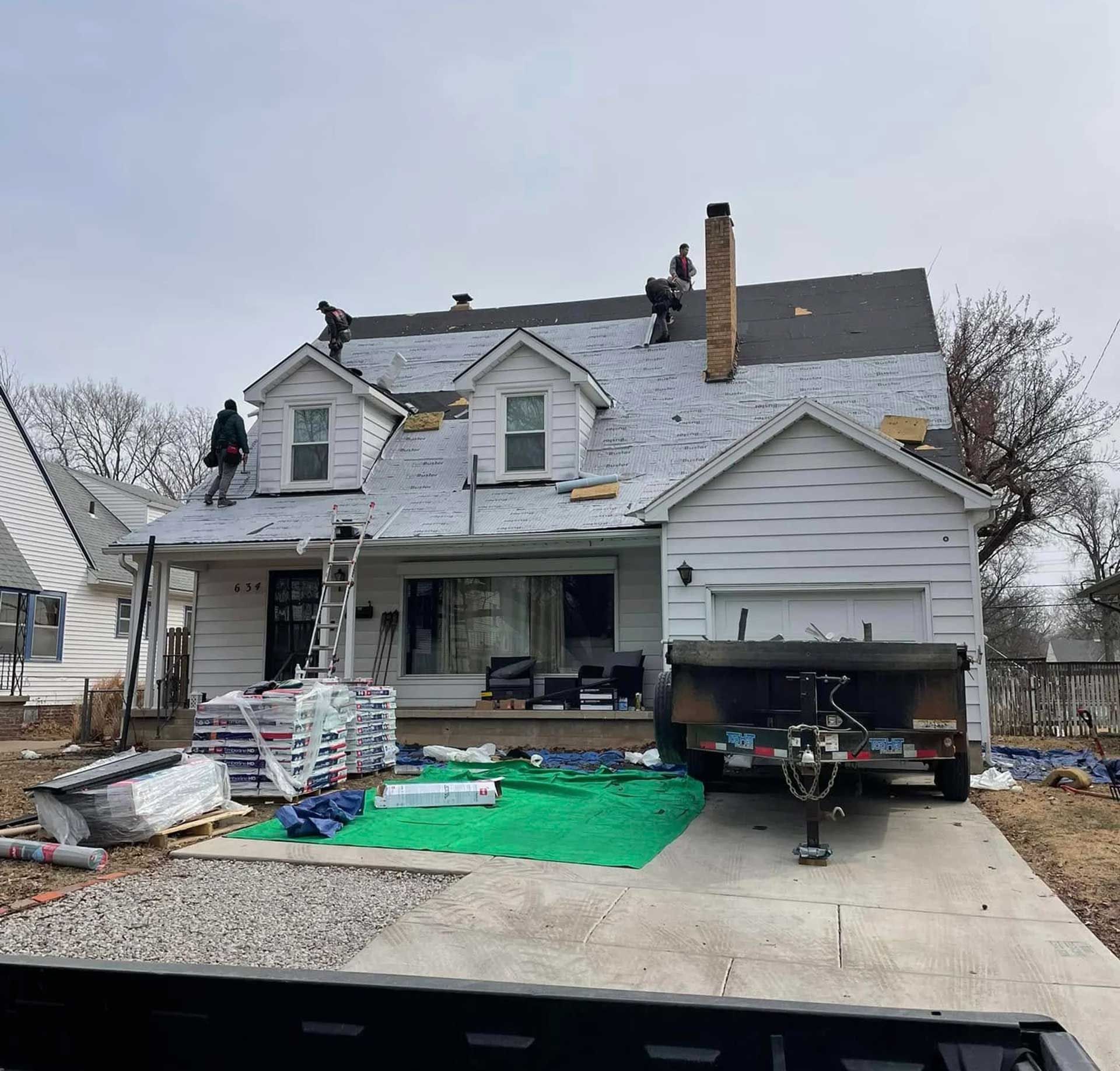 Roofers on a white house replacing the roof. Construction materials are on the ground.