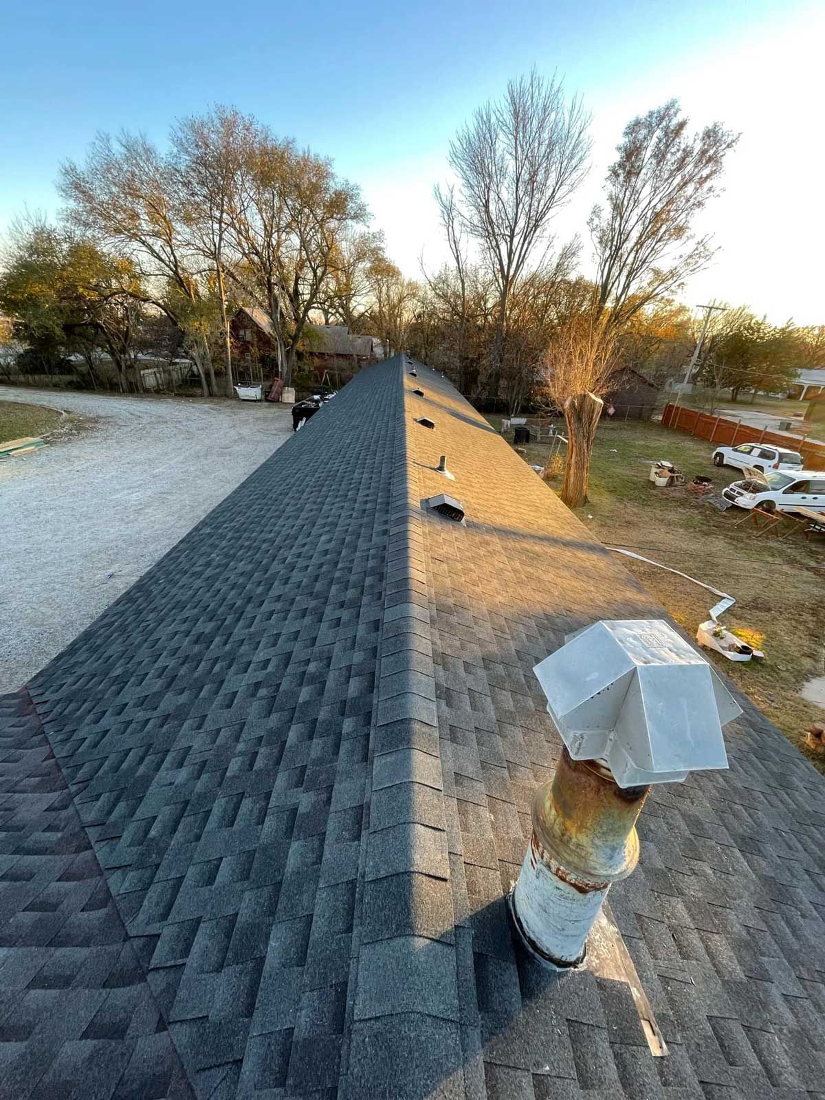 Rooftop view with dark asphalt shingles, exhaust vent in foreground, gravel driveway and trees in background.