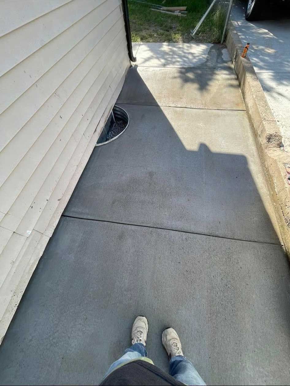 Person standing on a concrete walkway beside a beige-sided building, near a window well.