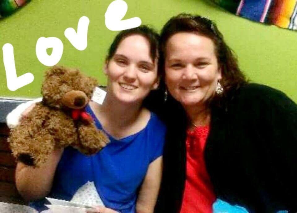 Two women are posing for a picture with a teddy bear in front of a wall that says love