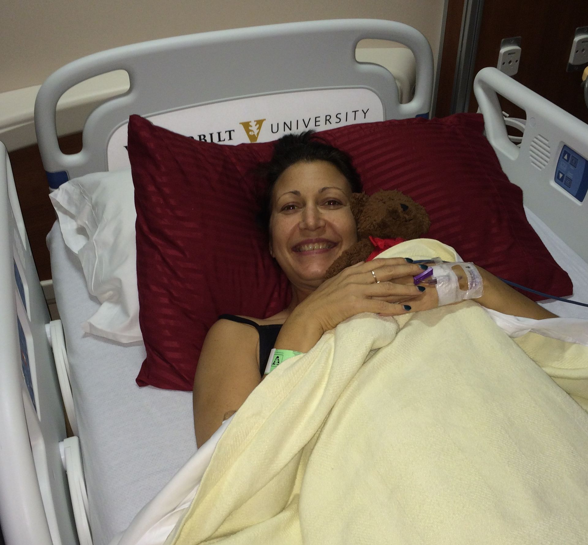 A woman is laying in a hospital bed holding a teddy bear and smiling