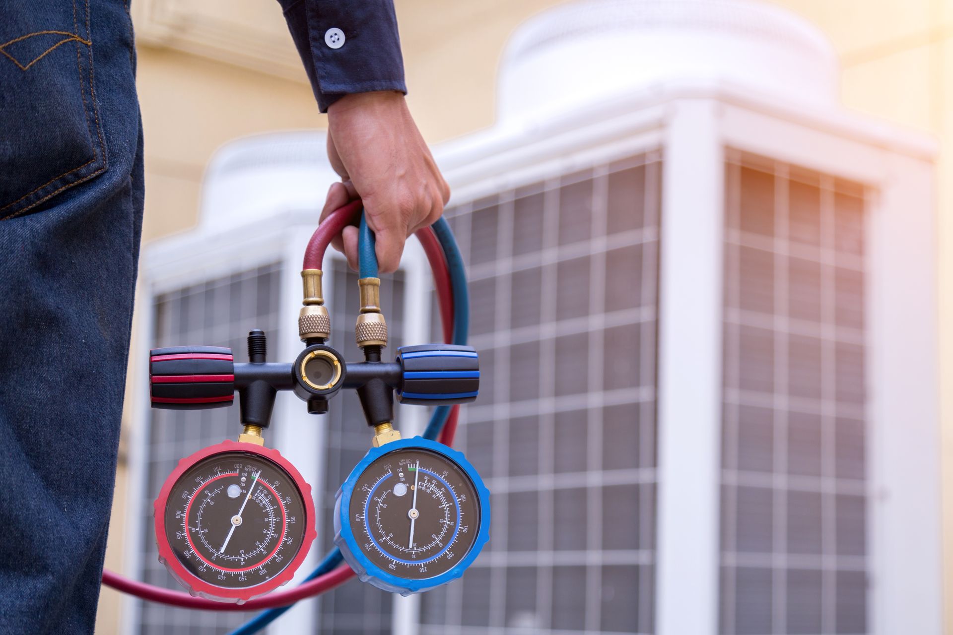 Person holding gauges, hoses in front of an AC unit. Red and blue gauges, jeans, sunlight.