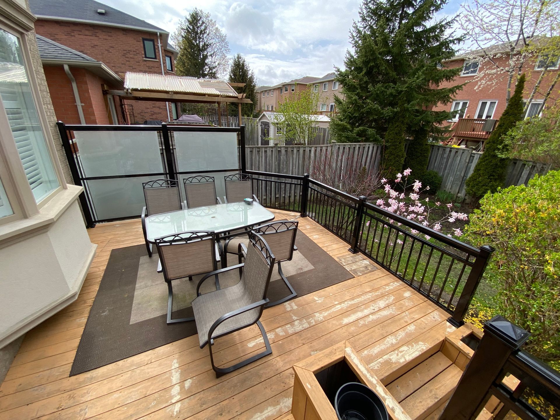 Outdoor wooden deck with glass-top dining table and privacy panels, surrounded by backyard garden.