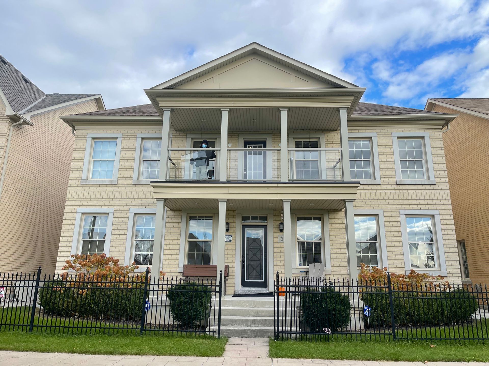 Two-story beige house with balcony, black fence, and bushes against a cloudy sky.