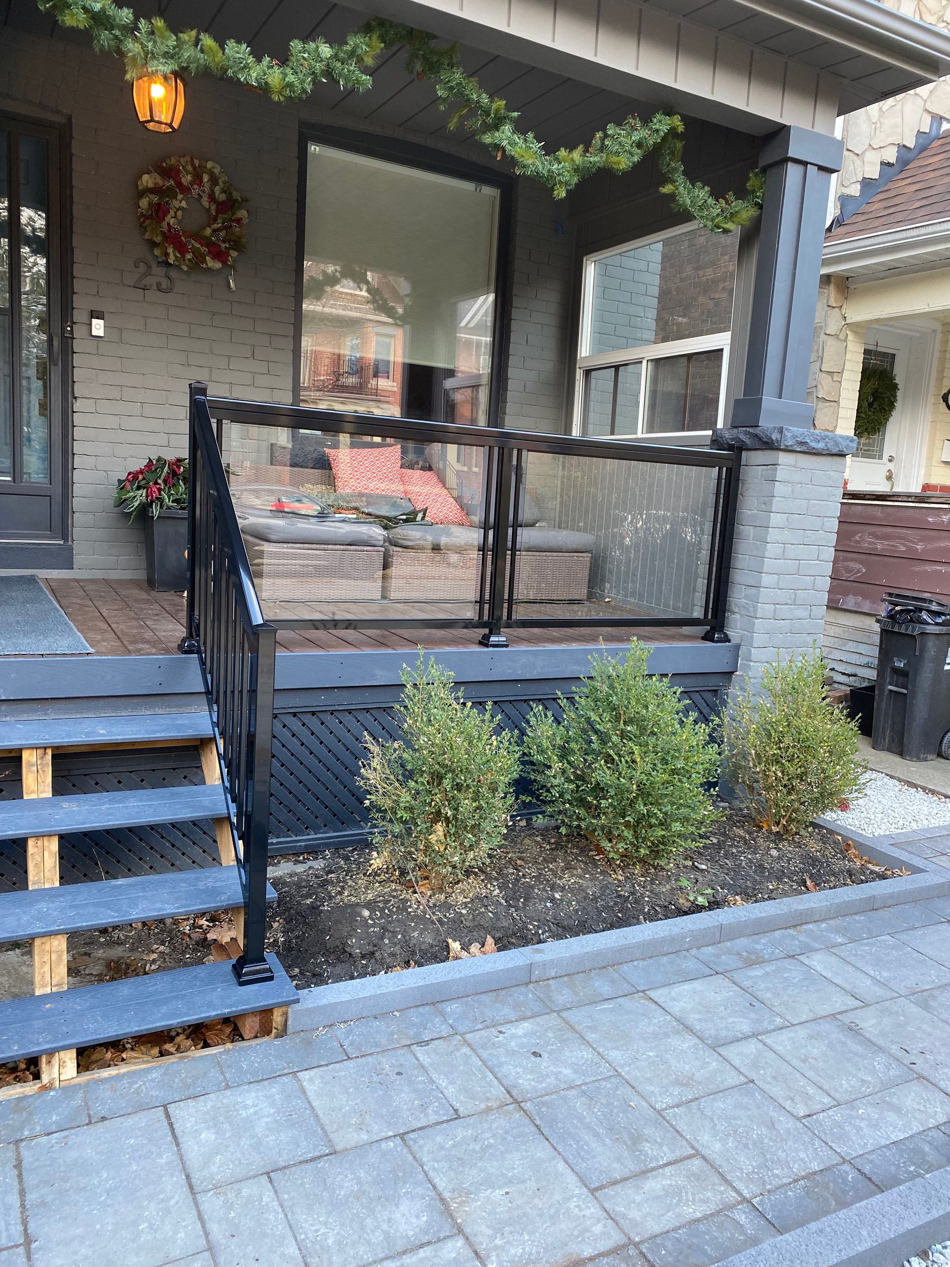 Front porch with glass railing, steps, and greenery. Black railing, grey stone, and house with Christmas decor.
