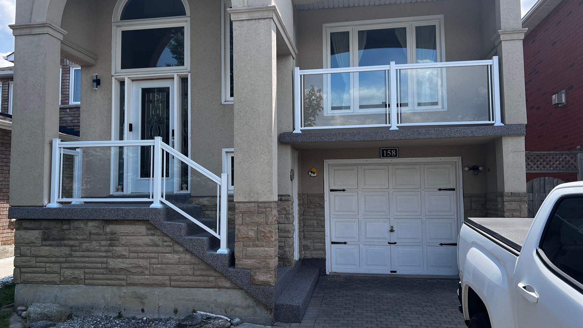 Two-story house with white trim, glass railings, beige exterior, and a white garage door. A white pickup truck is parked on the right.