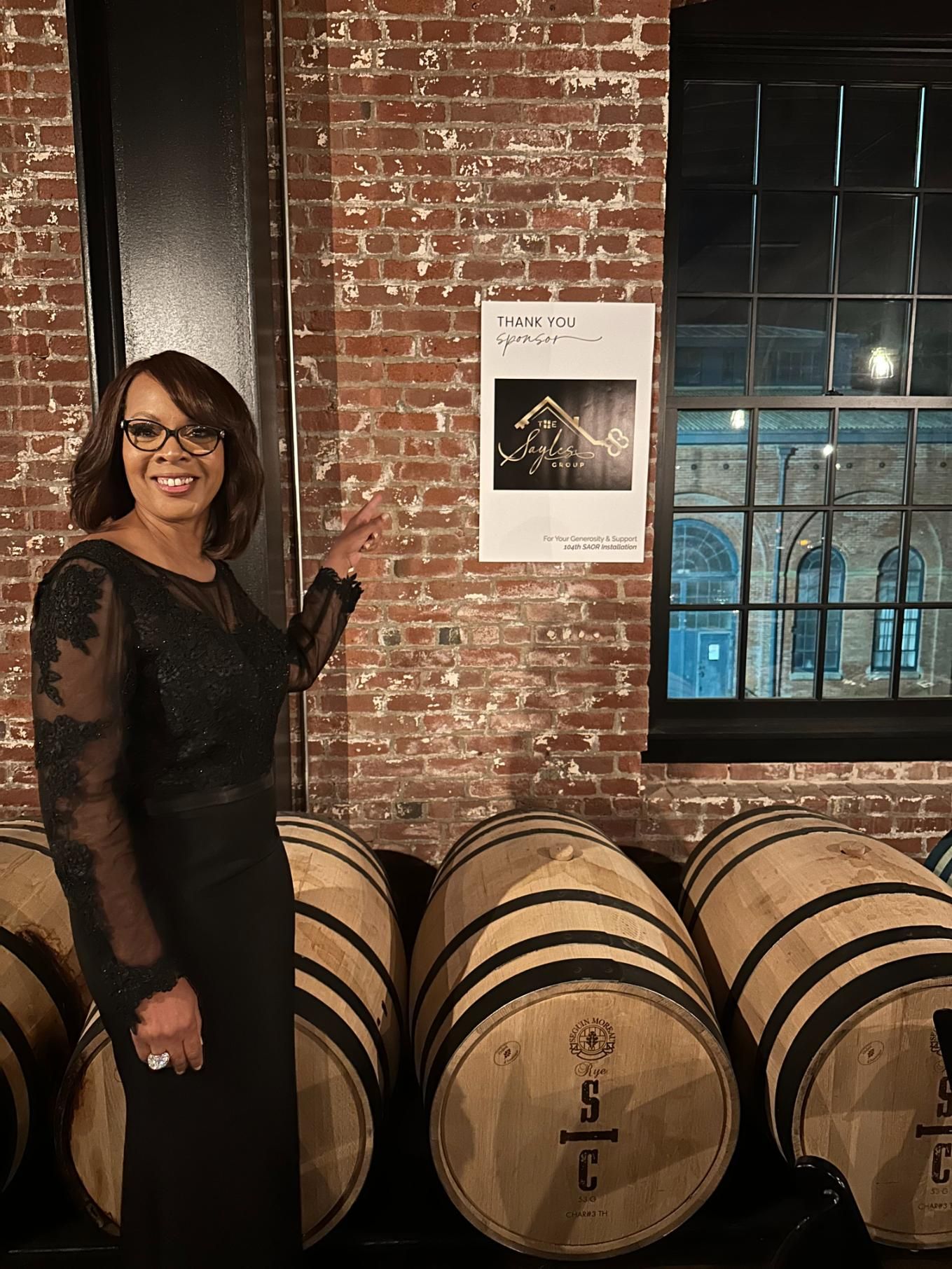A woman in a black dress is standing in front of a row of wine barrels.