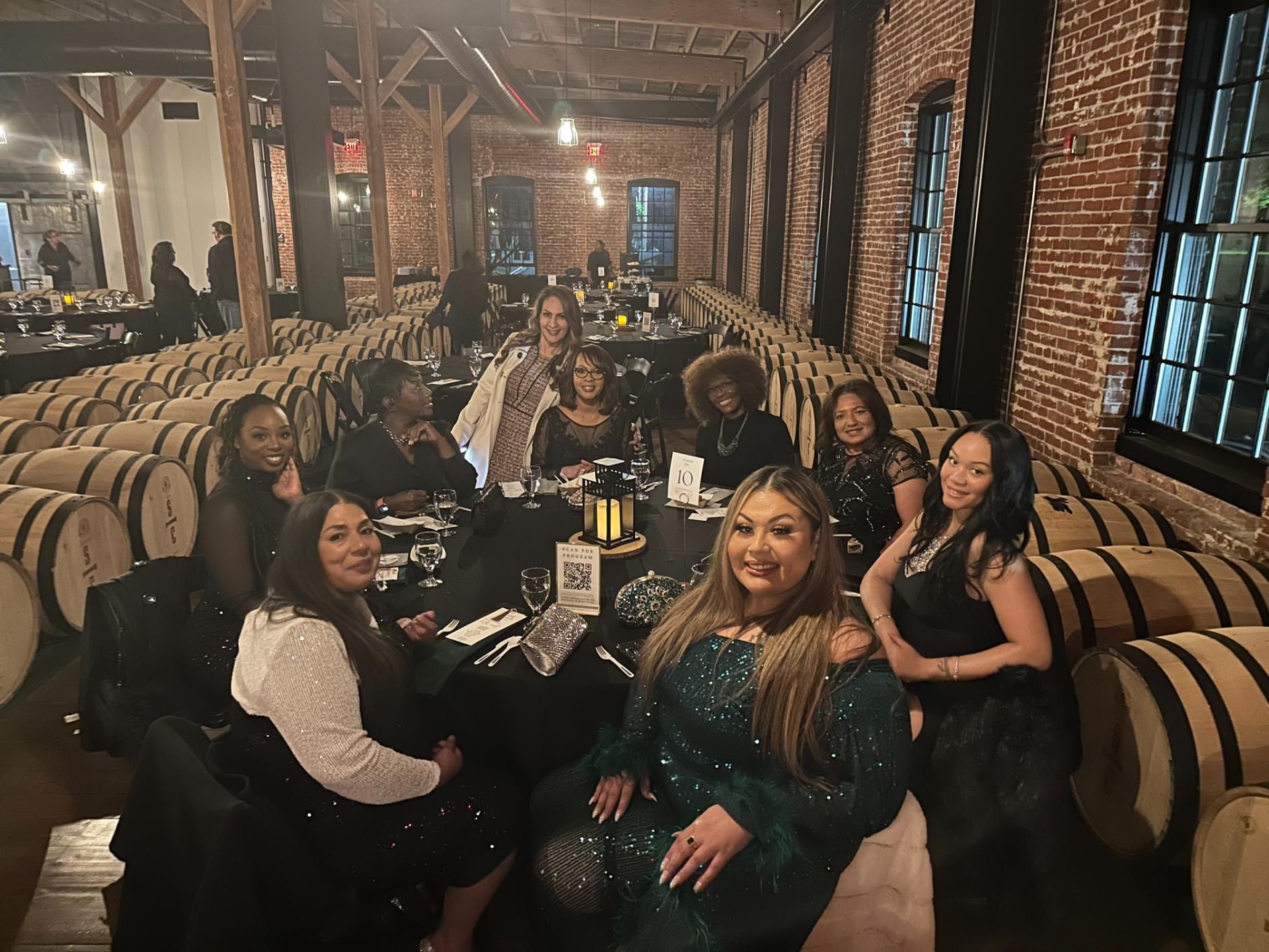 A group of women are sitting around a table in a room filled with barrels.