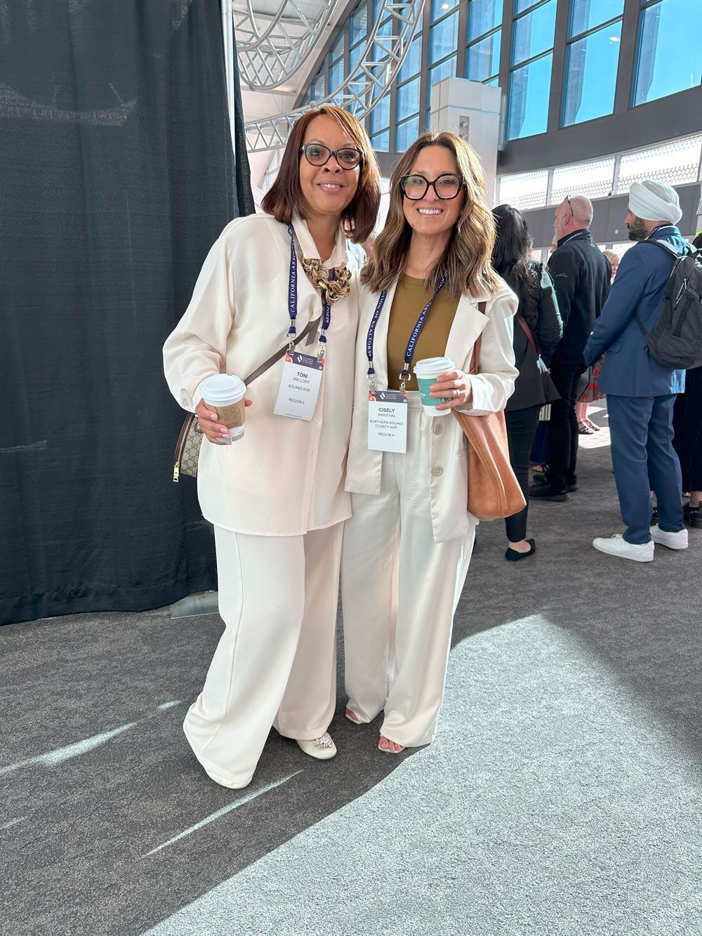 Two women are standing next to each other in a room holding cups of coffee.