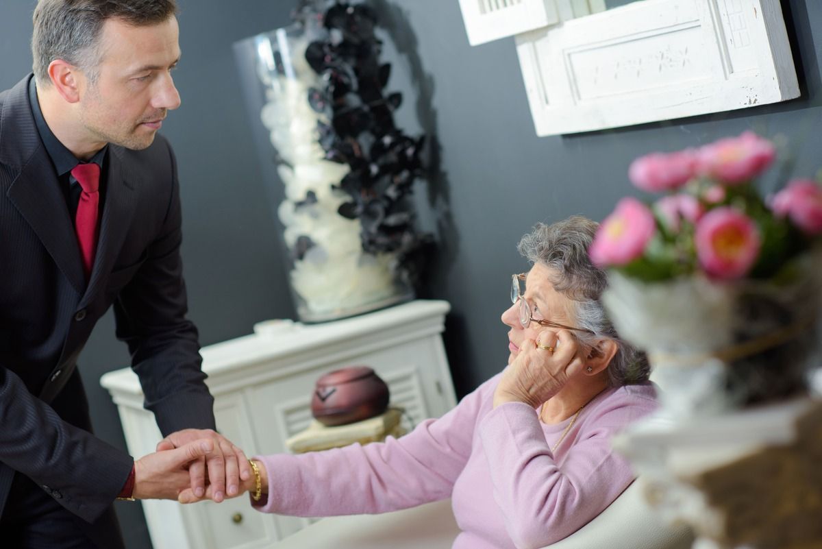Man in suit helping an elderly person. Man holds hand of seated woman. Interior, flowers and décor.