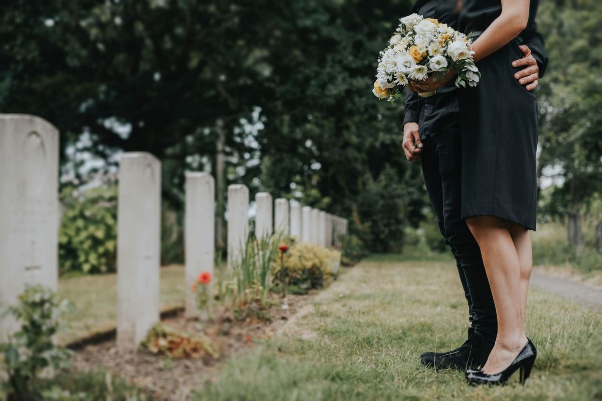 Couple standing in a cemetery, woman holding flowers, man has arm around her.
