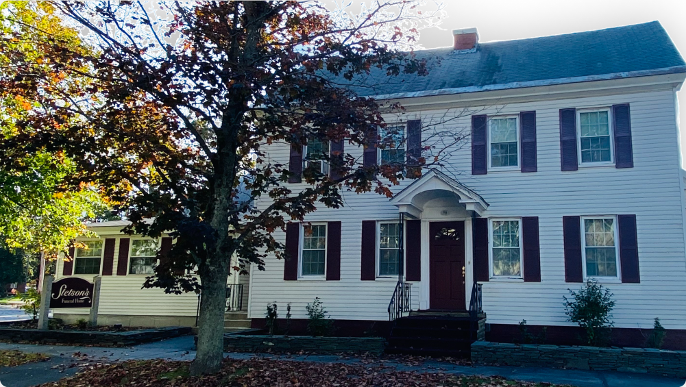 White two-story building with dark red shutters, a dark red door, and a sign that reads
