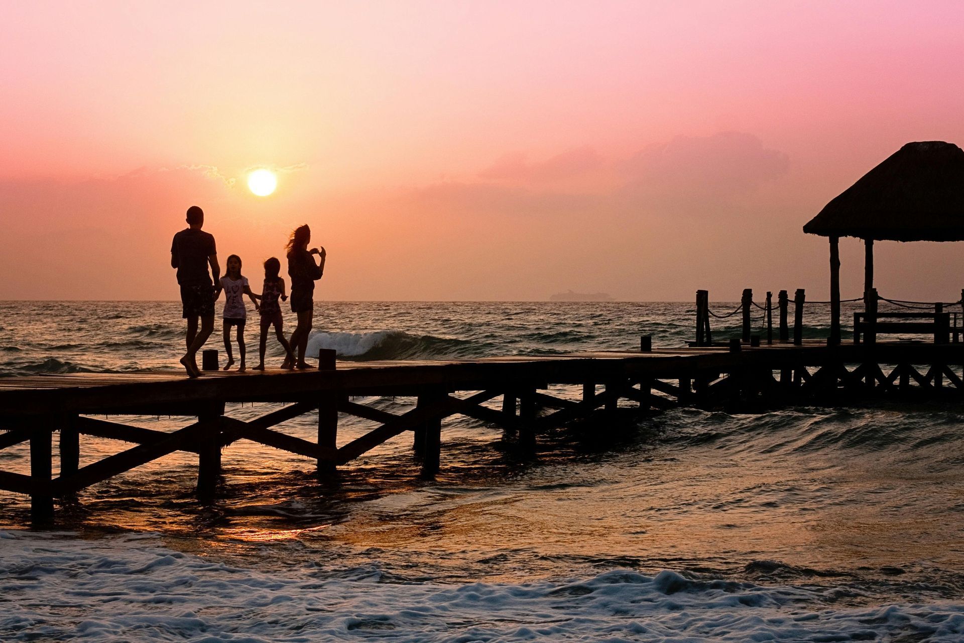 Family walking on a wooden pier at sunset with a small gazebo, ocean waves, and pink sky.