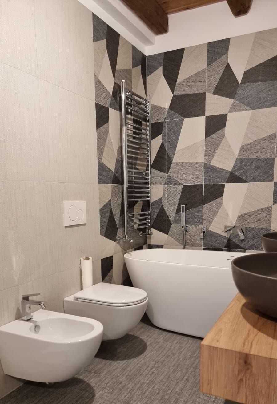 Bagno moderno con una parete decorativa a motivi geometrici, vasca bianca, WC sospeso, bidet e mobiletto in legno.
