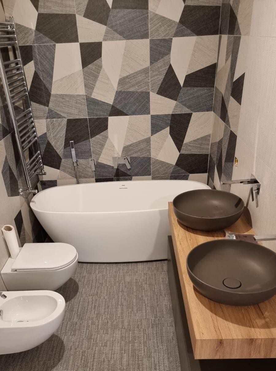 Bagno moderno con una parete decorativa a motivi geometrici, una vasca da bagno freestanding bianca, due lavabi a ciotola e un WC/bidet.