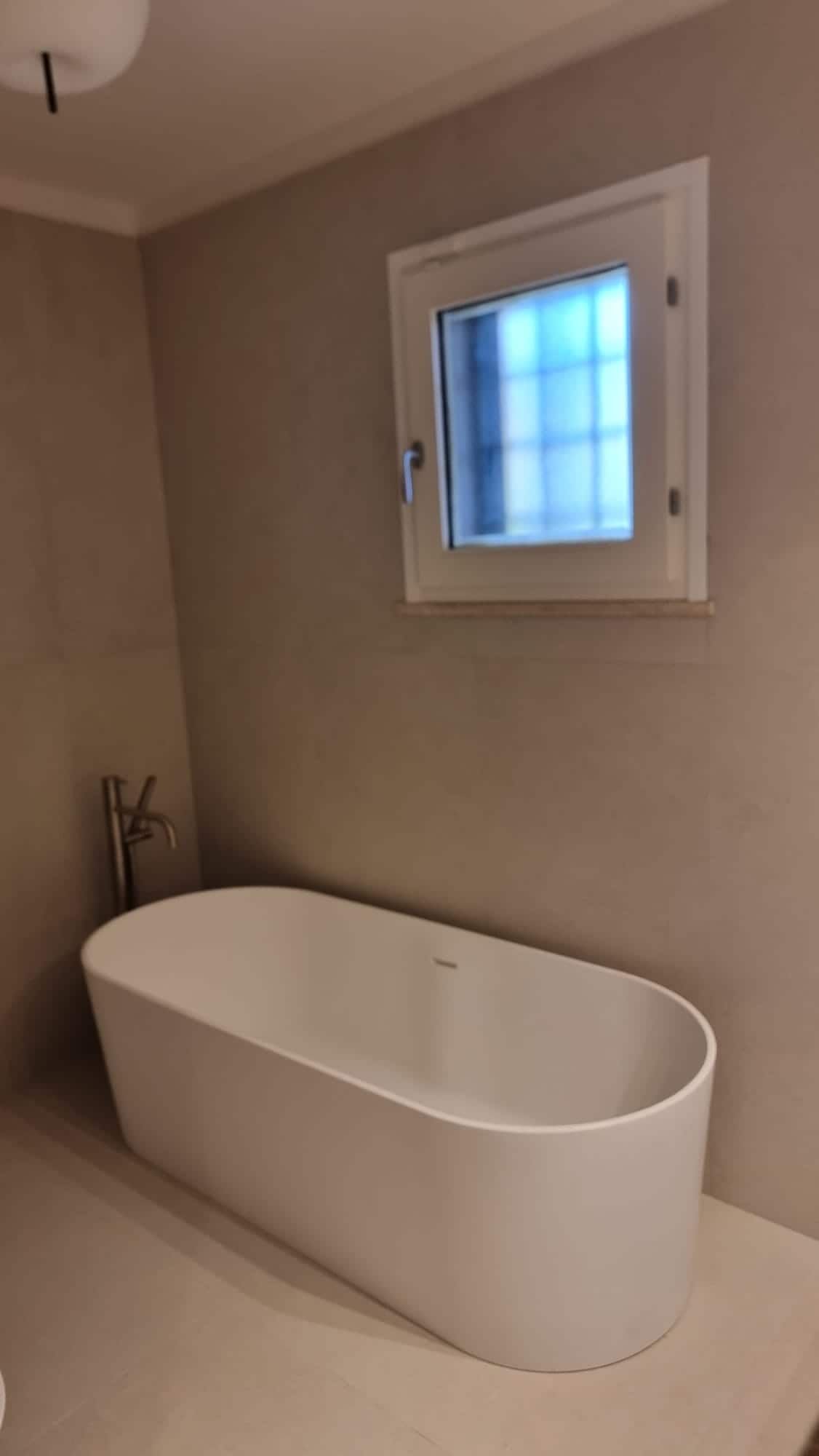 Una moderna vasca da bagno freestanding di colore bianco è addossata a una parete semplice e chiara, sotto una piccola finestra.