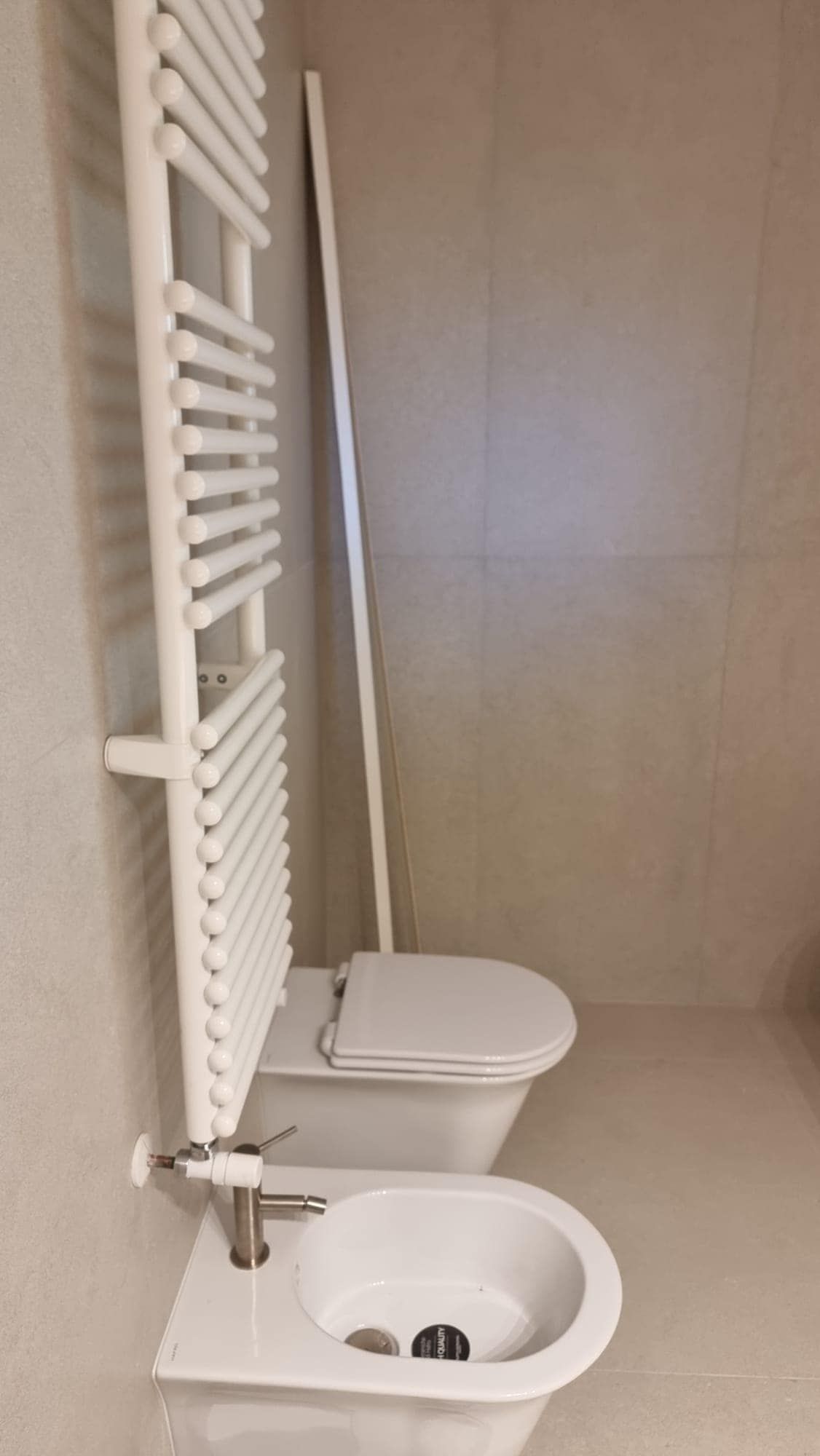 In un bagno dai colori neutri, un radiatore bianco a parete è sospeso sopra un bidet bianco e un WC sospeso.