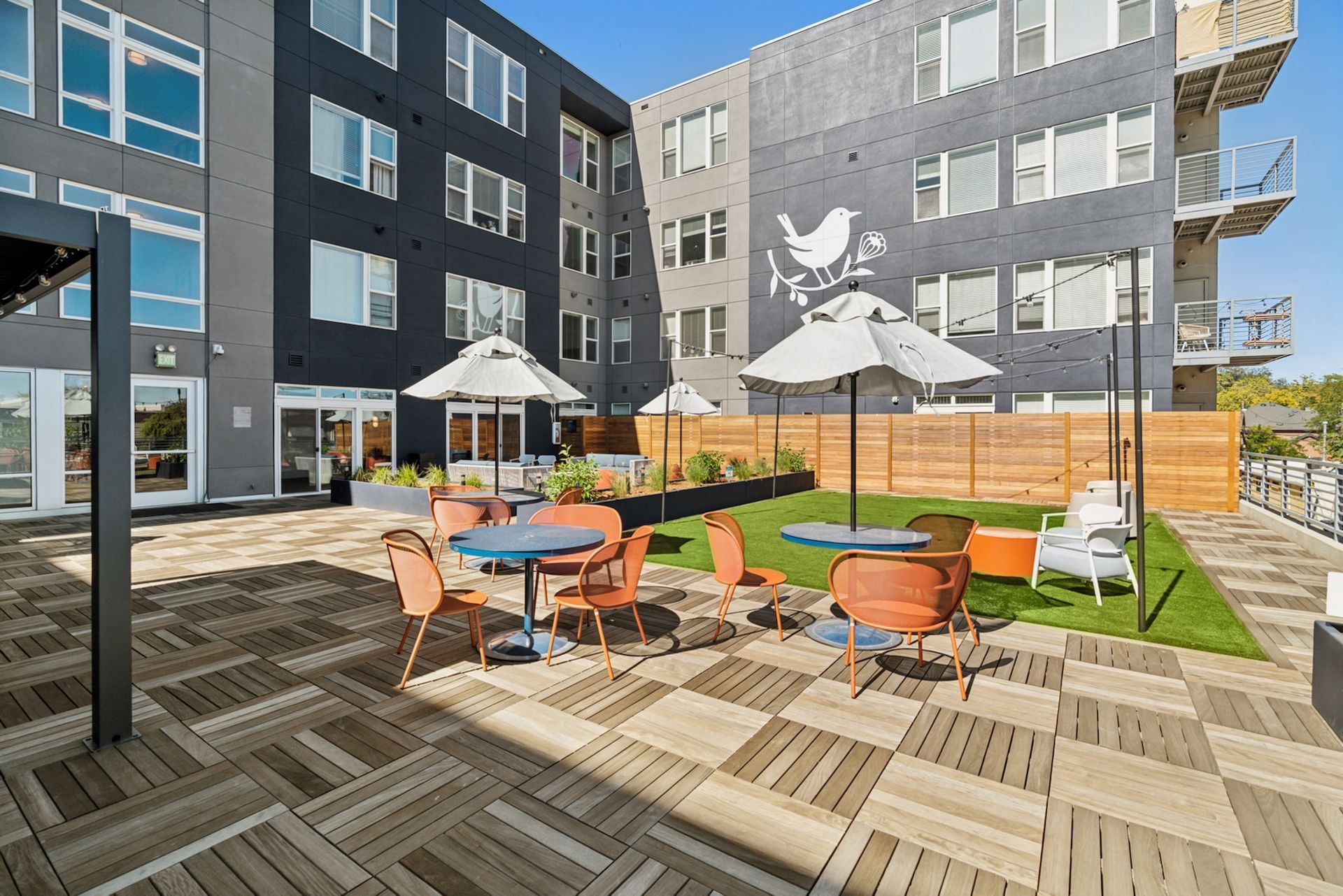 Outdoor patio with tables, chairs, and umbrellas.  Apartment building in background with a mural.