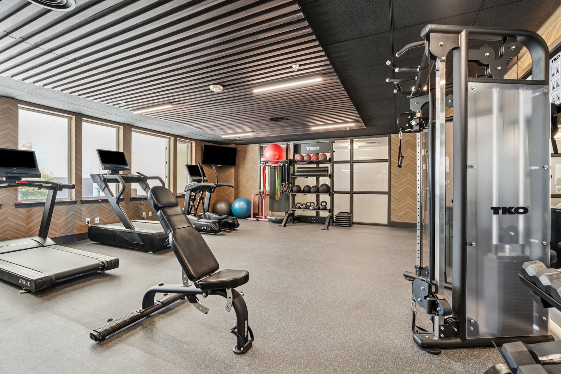 Gym interior: treadmills, weight bench, weights, and cable machine. Windows, gray floor, striped ceiling.
