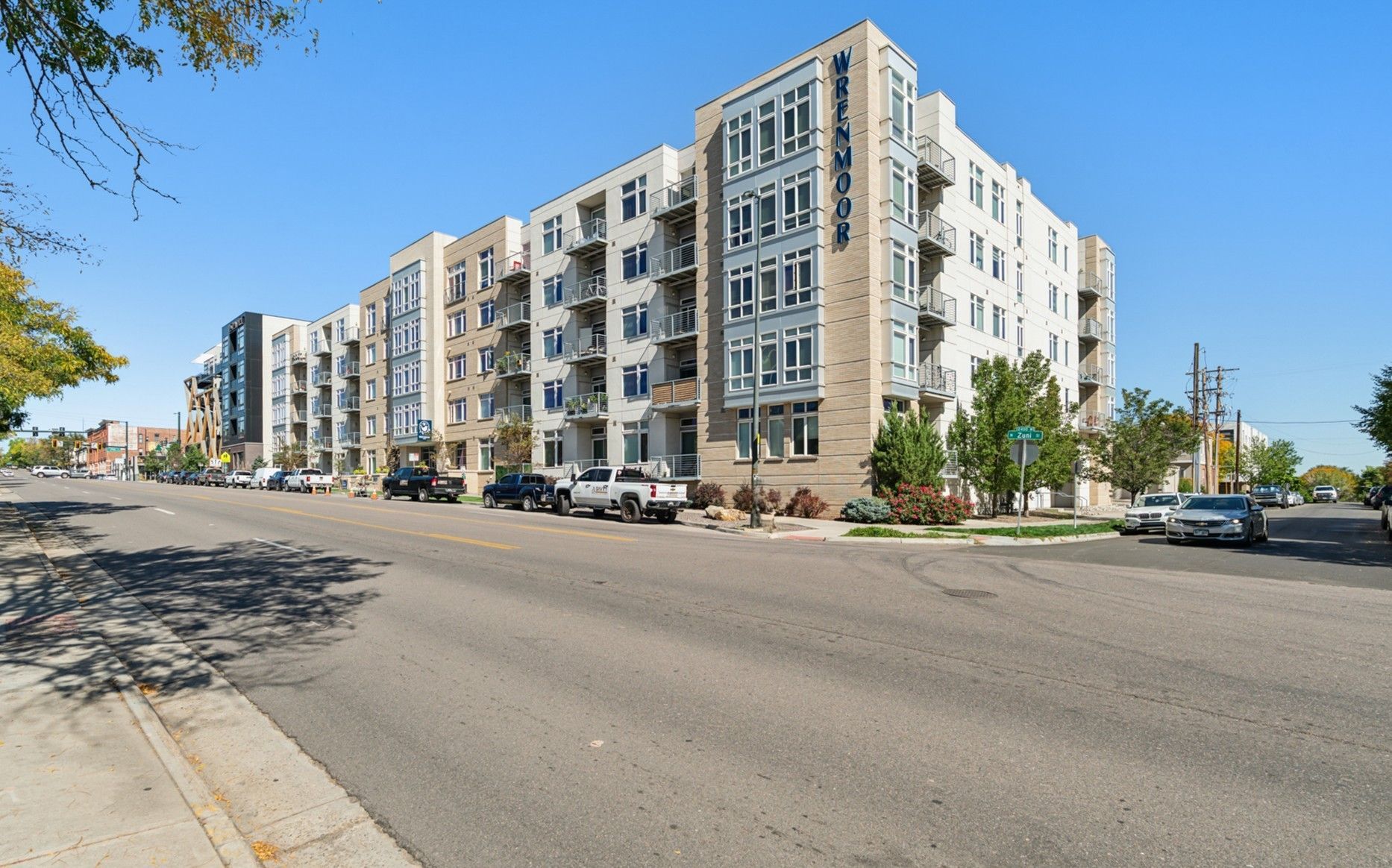 Multi-story apartment building on a city street. Bright blue sky. Several cars parked along the building.