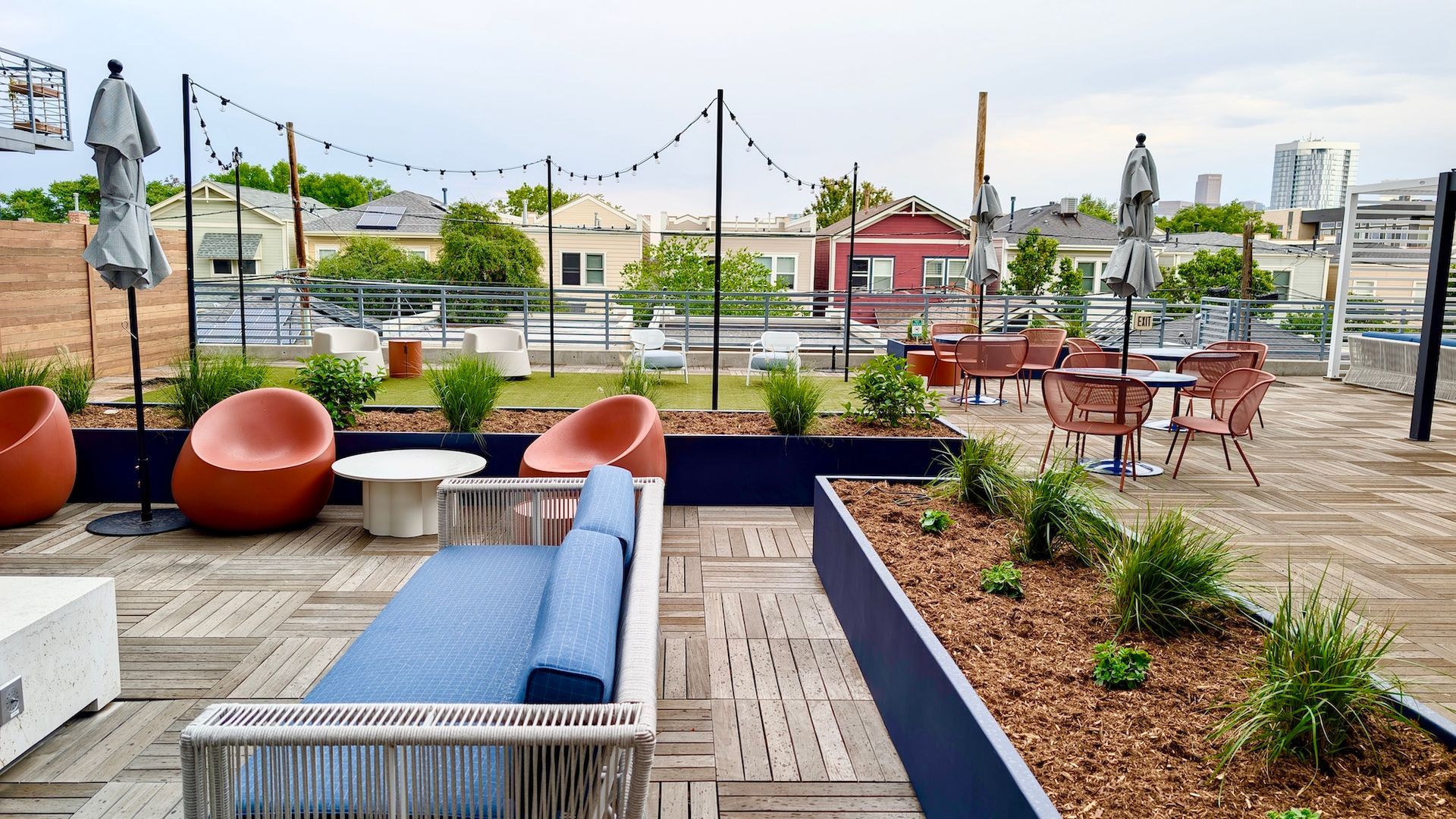 Rooftop patio with seating: orange chairs, blue couch, tables, string lights, plants, and city view.