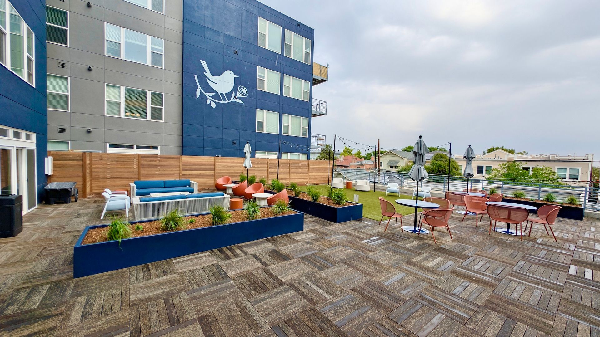 Rooftop patio with lounge seating, dining tables, and a blue building with a bird mural.