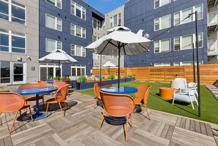 Outdoor patio with blue tables, orange chairs, umbrellas, and green turf, surrounded by a modern building with blue and gray walls.