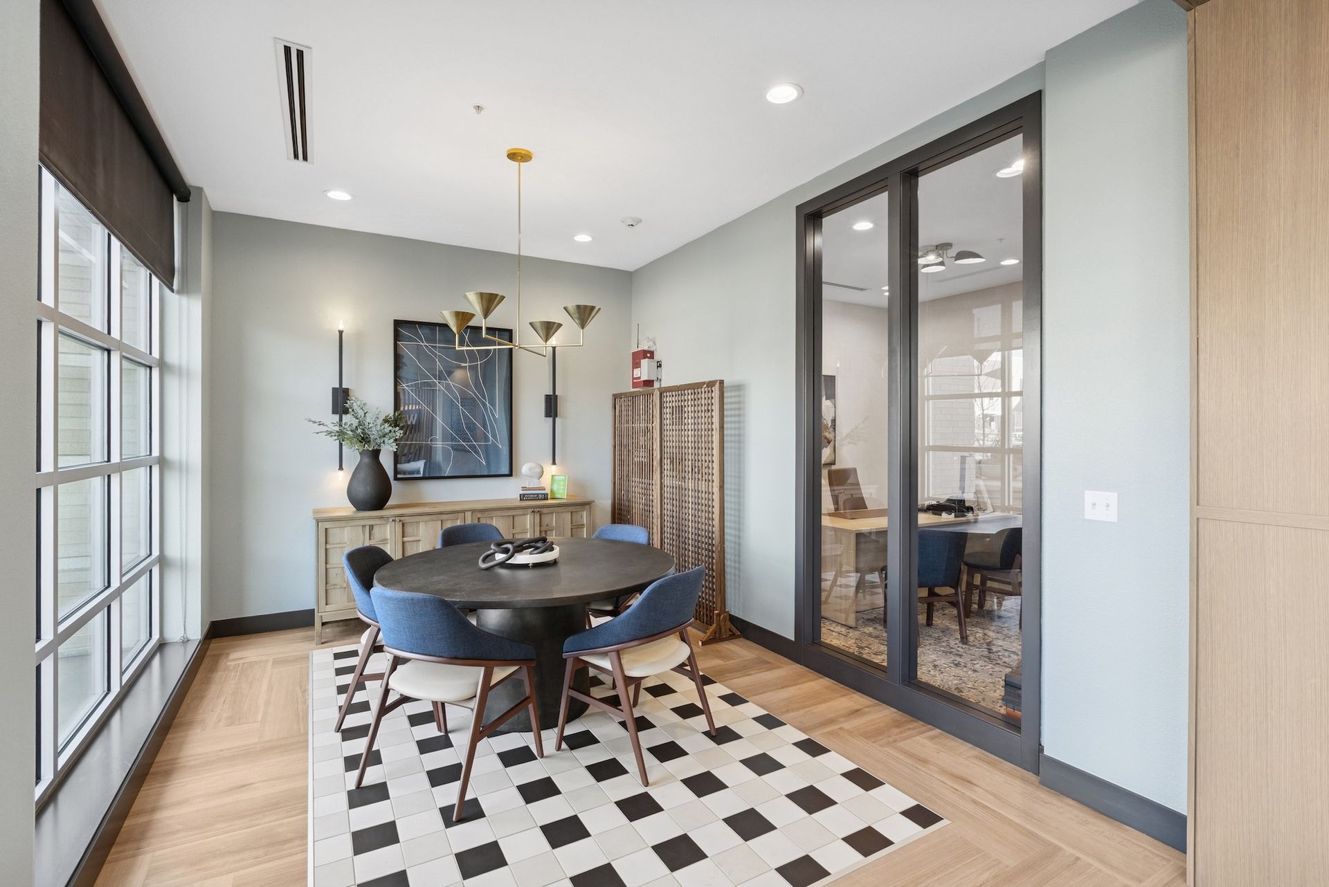 Dining room with round table, blue chairs, buffet, and patterned rug. Large windows and a glass door.