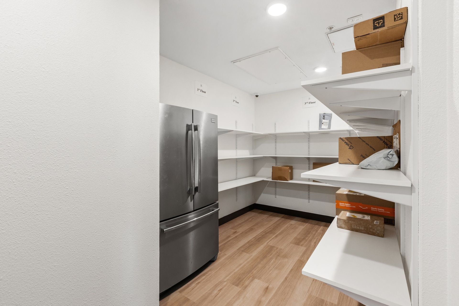 Walk-in pantry with white shelves, stainless steel refrigerator, and wood floor. Boxes and items are on shelves.