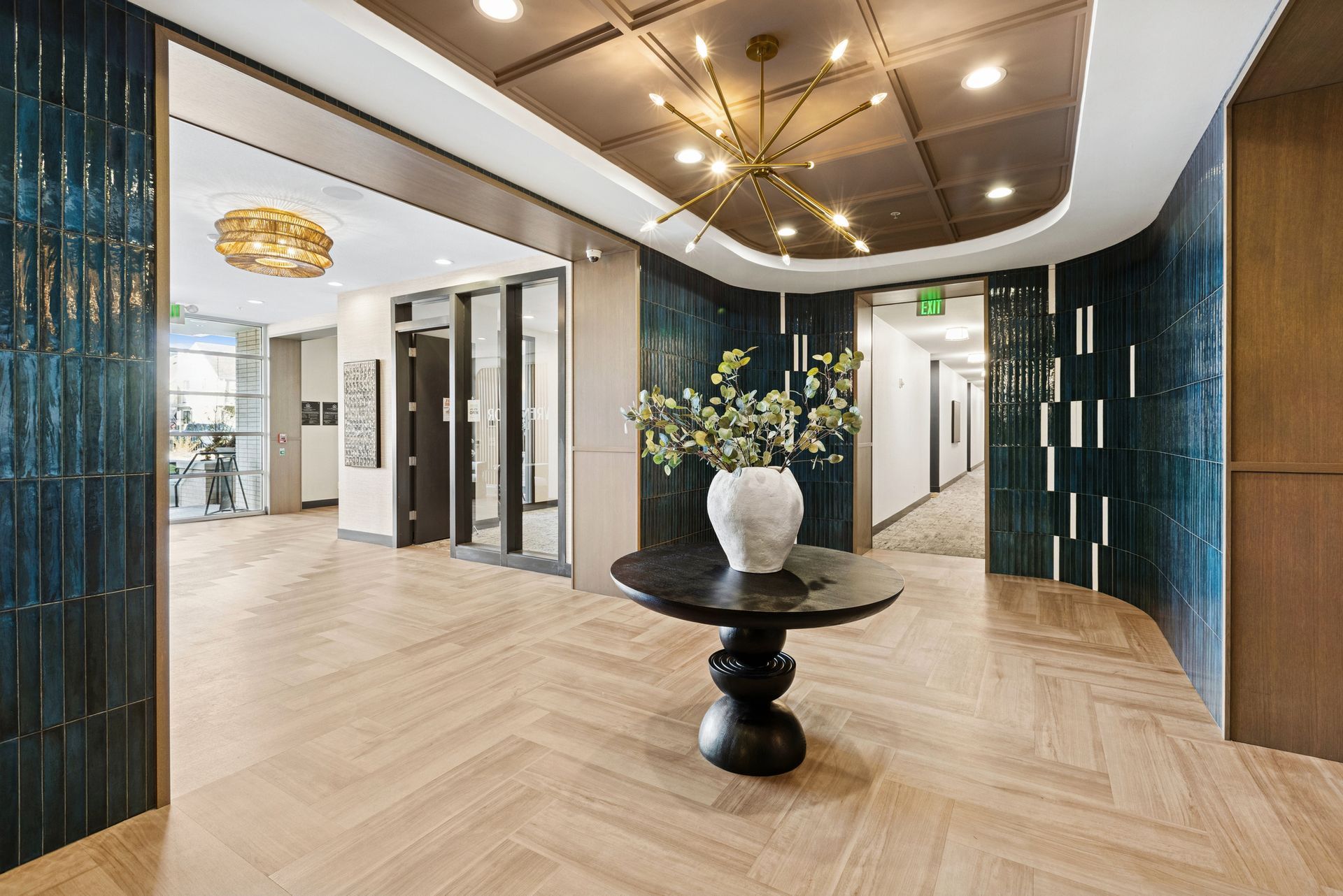 Elegant apartment lobby with teal tile accents, gold chandelier, dark wood table with flowers, and herringbone wood floor.