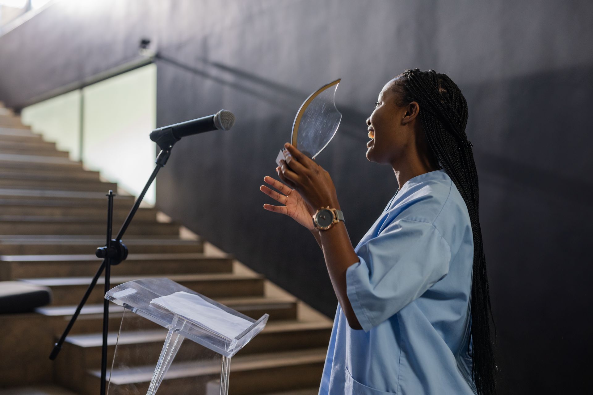 Woman speaking at a lectern, holding an award. Staircase and dark wall in the background.