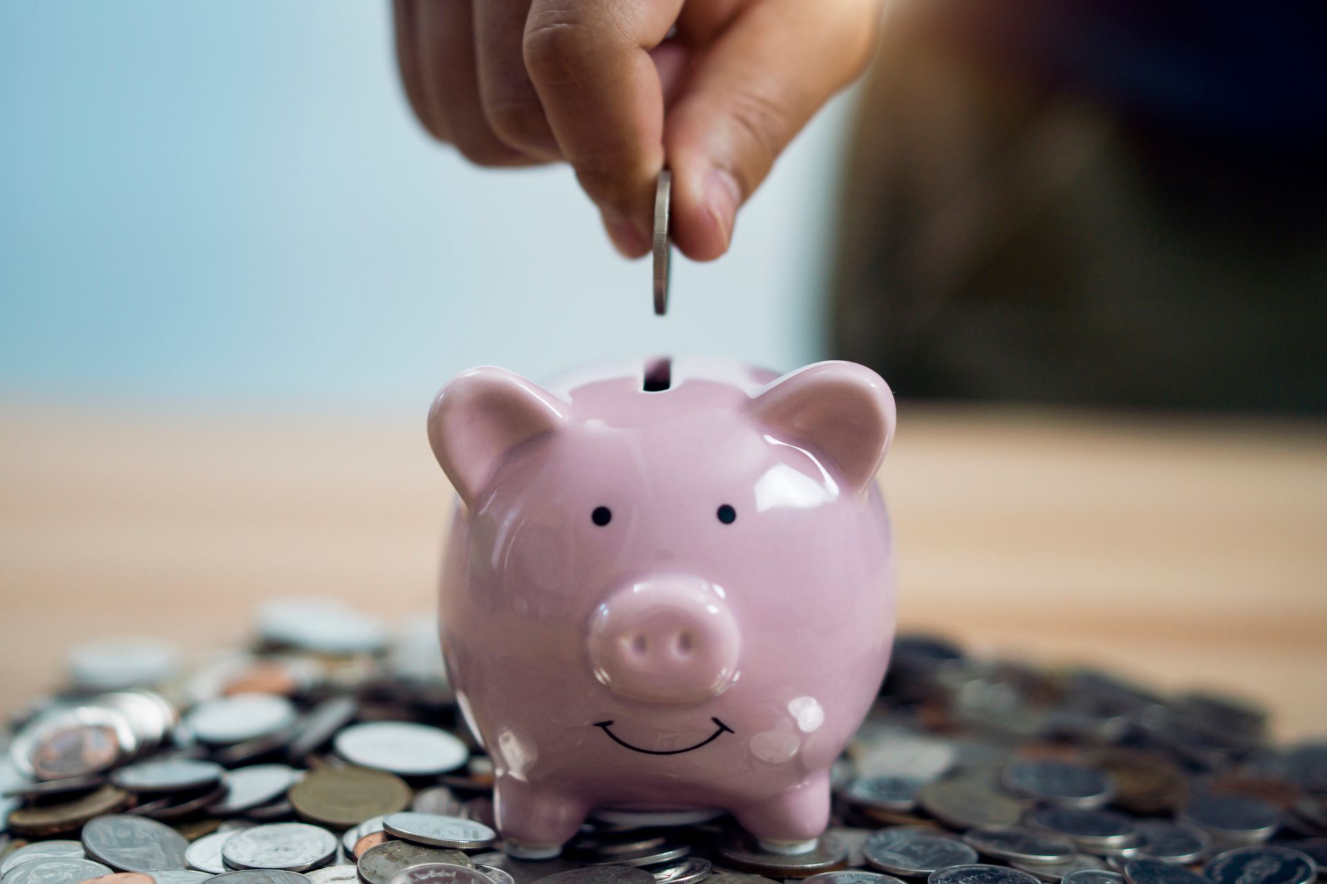 Person putting a coin into a pink piggy bank, surrounded by a pile of coins on a wooden table.