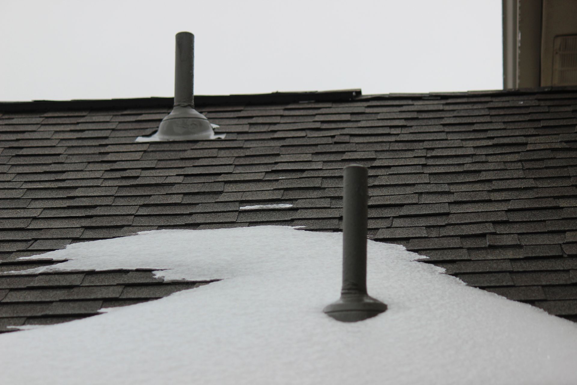 Snow-covered rooftop with two gray vent pipes. Dark asphalt shingles and a cloudy sky.